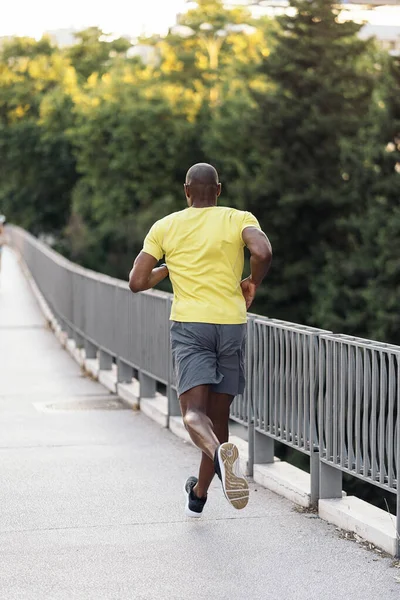 黄色のTシャツを着たアフリカ系アメリカ人スポーツマンの後ろ姿と 屋外の街の歩道をジョギングするショートパンツ — ストック写真