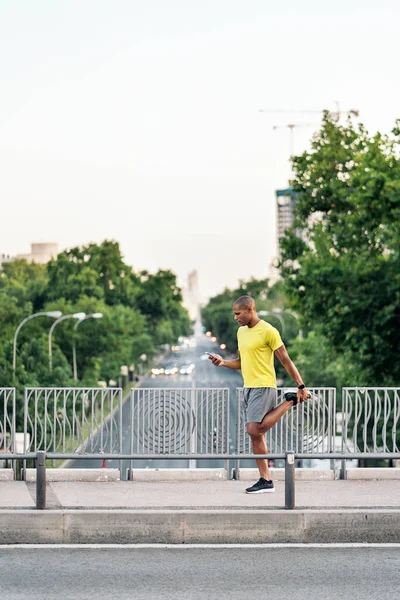 African American man stretching his left leg and holding phone in the footbridge at day.