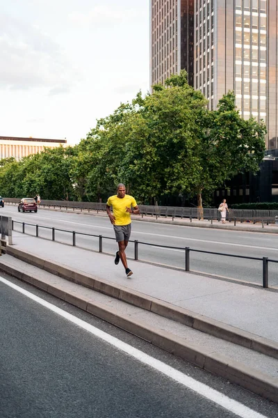 Front side view of an african American sportsman with yellow t-shirt and shorts jogging on sidewalk in middle in the city.