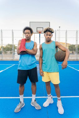 Basketbol topu tutan, gülümseyen ve basketbol sahasında kameraya bakan havalı Afro-Amerikan erkekleri..