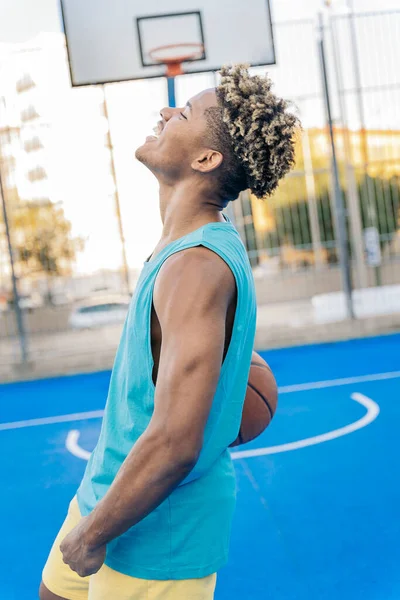 Happy afro american basketball player looking at camera holding the ball under his arm in a street basketball court.