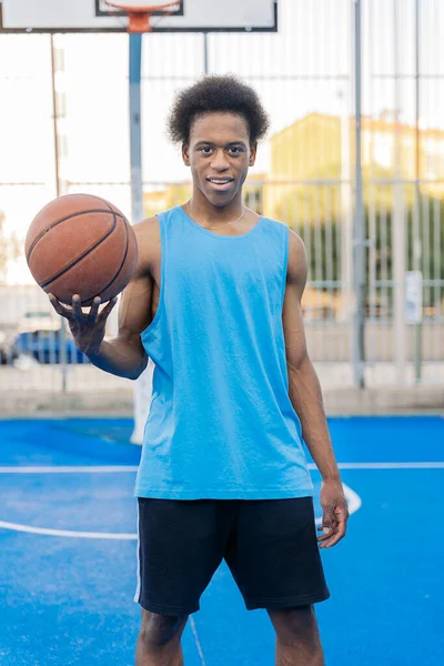 Happy afro american basketball player looking at camera holding the ball with his hand in a street basketball court.