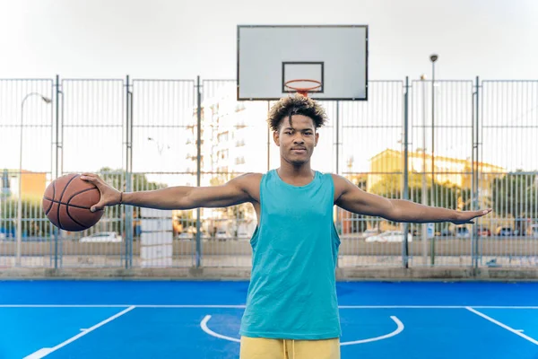 Happy afro american basketball player looking at camera holding the ball with one hand in a street basketball court.