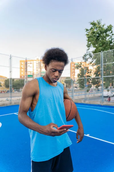 ストリートバスケットボールコートで彼の腕の下にボールを保持しながら 携帯電話を使用して若いアフロアメリカのバスケットボール選手 — ストック写真
