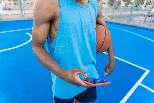 ストリートバスケットボールコートで彼の腕の下にボールを保持しながら 携帯電話を使用して若いアフロアメリカのバスケットボール選手 — ストック写真
