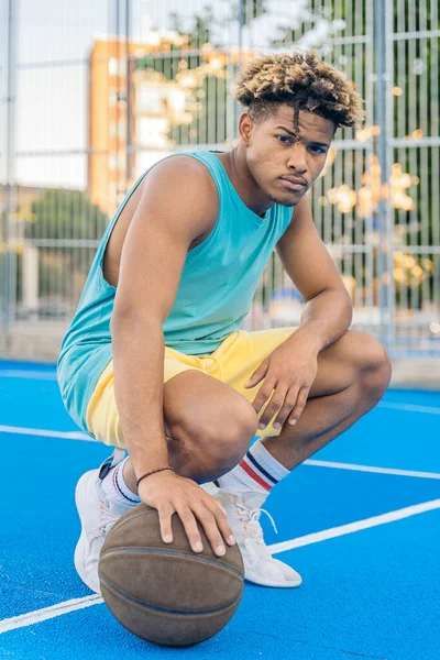 Portrait of an afro american basketball player crouching and holding the ball on the ground looking seriously at camera on the court.