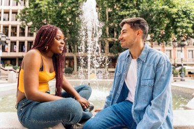 Çok kültürlü genç arkadaşlar çeşmenin yanında oturup konuşuyorlar ve birbirlerine bakıyorlar..