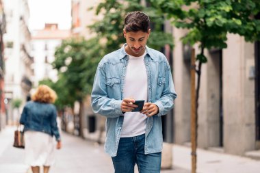 Yakışıklı, sıradan kıyafetler giyen, telefonunu kullanan ve sokakta yürüyen bir genç..