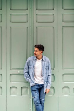 Havalı genç adam sıradan kıyafetler giyiyor gülümsüyor ve yeşil duvara bakıyor..