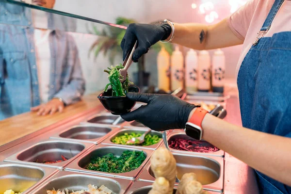 未被认可的餐馆员工戴着防护手套 将配料放入装有米饭和蔬菜的锅中 — 图库照片
