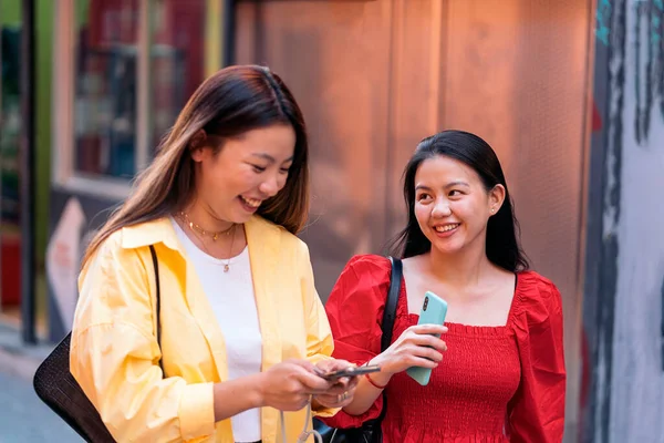 Young and pretty asian female friends laughing and using their phones in the street.