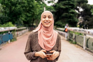 Parkta yürüyen ve cep telefonuyla yazan güzel Müslüman kız. Gülümsüyor ve kameraya bakıyor..