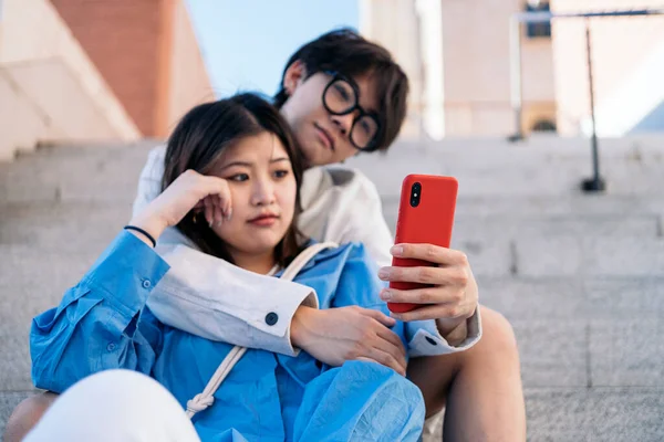 幸せなアジアのカップル一緒に通りに座って 彼らのスマートフォンを見て — ストック写真