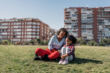 Annesi gülümseyip kameraya bakarken çimenlerde oturan tatlı afro kız..