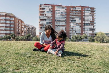 Saçları örgülü güzel siyahi bir kadın genç kızıyla çimenlerde oturuyor ve güneşli bir günün tadını çıkarıyor..