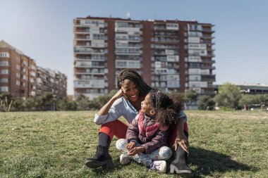 Saçları örgülü güzel siyahi bir kadın genç kızıyla çimenlerde oturuyor ve güneşli bir günün tadını çıkarıyor..