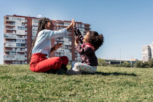 愛らしいですアフロ女の子で草の中に座って彼女の母親と楽しいです日中晴れた日 — ストック写真