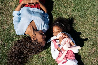 Saçları örgülü siyahi bir kadın. Küçük kızının yanına uzanmış ve gülümsüyor..