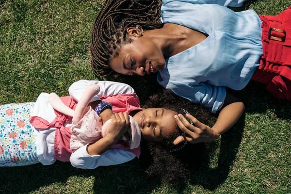 Adorable afro girl hugging stuffed animal lying down in the grass with her mother. They are smiling and having fun.