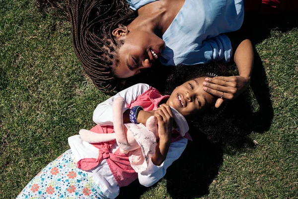 Adorable afro girl hugging stuffed animal lying down in the grass with her mother. They are smiling and having fun.