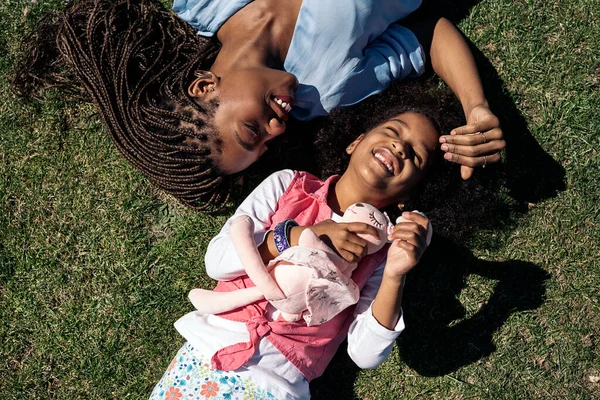 Adorable afro girl hugging stuffed animal lying down in the grass with her mother. They are smiling and having fun.