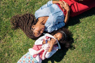 Saçları örgülü siyahi bir kadın. Küçük kızının yanına uzanmış ve gülümsüyor..