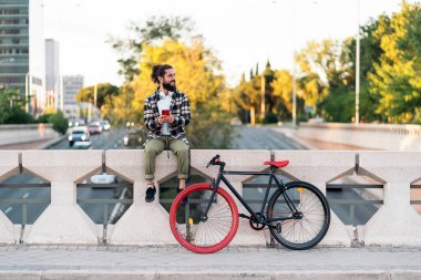 Şehirde bisikletinin yanında oturan ve cep telefonunu kullanan sakallı havalı bir adam..