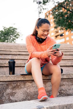 Spor yaptıktan sonra cep telefonunu kullanan genç bir kadın. Spor kıyafetleri ve kulaklık takıyor..