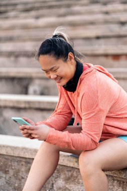 Spor yaptıktan sonra cep telefonunu kullanan genç bir kadın. Spor kıyafetleri ve kulaklık takıyor..