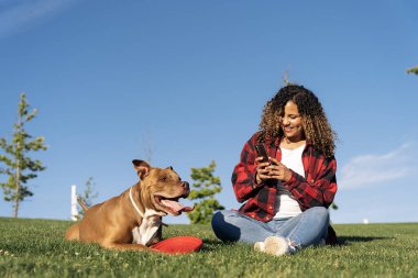 Afrikalı bir kadın köpeğiyle parkta oturuyor ve cep telefonunu fotoğraf çekmek için kullanıyor..