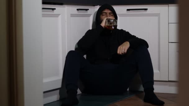 フード付きの放置された酔った男がキッチンフロアに座り ボトルからウィスキーを飲みます うつ病アルコール依存症孤独の概念 — ストック動画