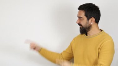 Sıradan kıyafetler giymiş sakallı bir adam gülümseyip kameraya bakıyor ve iki eliyle ve parmaklarıyla kenarı işaret ediyor..