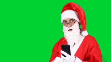 Noel Baba ayakta duran akıllı telefon görünümlü mutlu bir galibiyet, yeşil arka planda izole edilmiş bir jest yapıyor. Noel bir tatil konseptidir..