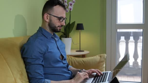 一个留着胡子的男人坐在沙发上与笔记本电脑一起工作 聪明的工作概念 — 图库视频影像