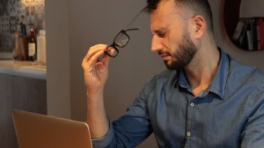 Sakallı adam bilgisayarla çalışıyor. Evde oturup dizüstü bilgisayarda yazma yorgunluğu, gözlükleri çıkarmak, burnu ve gözleri ovuşturmak yorgunluk ve baş ağrısı hissetmek. Stres ve hüsran konsepti