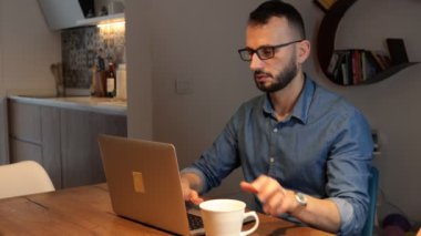 Sakallı ve gözlüklü bir adam laptopuyla evde oturup çay ya da fincandan kahve içiyor..