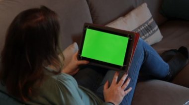 Evdeki kanepede oturan kız elinde yeşil ekranlı bir tablet tutuyor. Eğlence içeriği ya da öğrenme.  