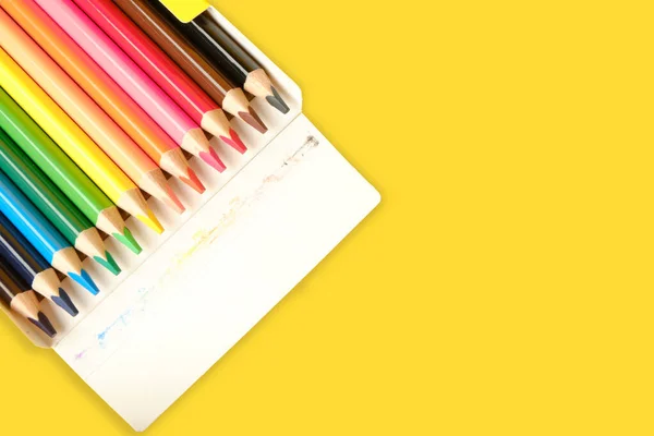 Multi-colored pencils for drawing in a box on yellow background with place for text.