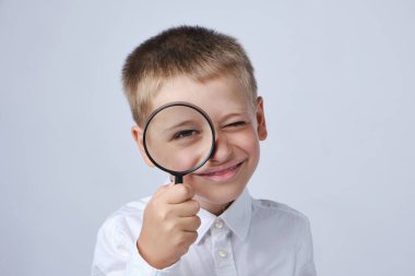 Smiling child looks at the camera through a magnifying glass on a gray background. Close-up.