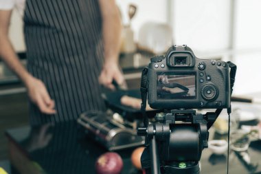 Kahverengi önlüklü adamın ocakta yemek pişirirken ve DSLR kamerasını tripoda çekerken yakın plan görüntüsü..