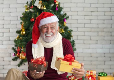Süslü Noel ağacının önünde elinde Noel hediyeleri tutan kırmızı süveter, eşarp ve Noel Baba şapkalı gülümseyen beyaz son sınıf öğrencisinin yakın plan portresi. İyi tatiller..  