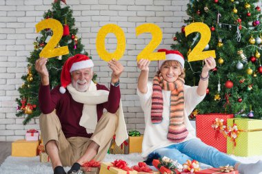 2022 yılının sarı parıltılı yılbaşı numaralarını taşıyan iki mutlu ve neşeli yetişkin evde yeni yılı, yeni yılı ve Noel 'i kutluyorlar. Oturma odasında süslü Noel ağacı. 