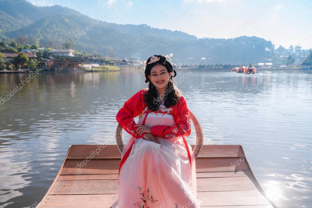 attractive Asian woman tourist wearing red Hanfu ancient Chinese traditional ethnic dress sitting on wooden boat at reservoir of Ban Rak Thai village, Mae Hong Son province, Thailand