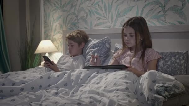 Дети, использующие цифровые устройства в постели — стоковое видео