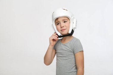 Koruyucu kasklı, acı çeken bir görünüşü olan küçük bir çocuk açık gri bir arka planda izole edilmiş tokayı düzeltir..