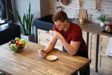 Gündelik giysiler içinde genç çekici bir adam mutfakta bir fincan kahve ve rahat bir evde bir telefonla oturuyor..