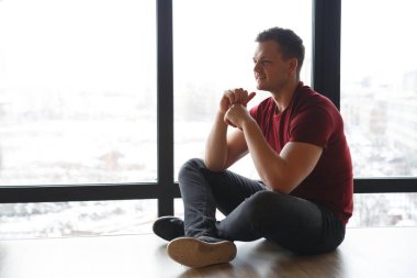 Kış günü panoramik bir pencerenin arka planında yerde oturan atletik bir adamın silüeti..