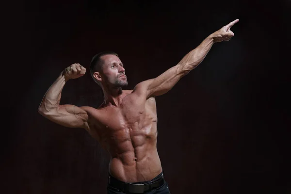 一个身材苗条 体格苗条 身体赤裸的运动员 他的一只手在黑暗的背景下指向右上身 表现出肌肉 — 图库照片