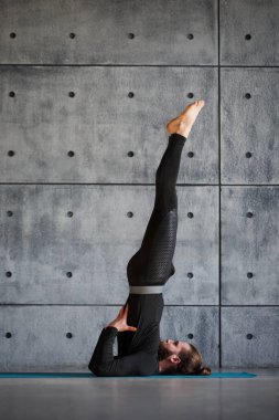 Siyah spor kıyafetli genç sakallı bir adam gri bir duvarda yoga yapıyor..