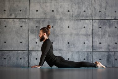 Siyah spor kıyafetli genç sakallı bir adam gri bir duvarda yoga yapıyor..